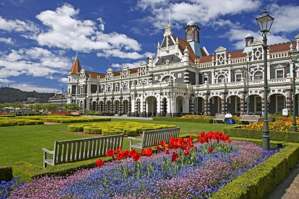 Entdecken Sie den historischen Bahnhof in Dunedin, Neuseeland, umgeben von bunten Blumen und einer einladenden Parklandschaft.